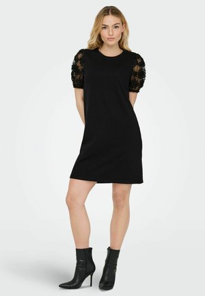 Robe noire à manches courtes avec des manches en dentelle florale bouffantes, encolure ronde et ourlet au genou. Associée à des bottines noires.