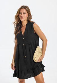 Robe noire sans manches avec un col en V, boutonnage sur le devant et ourlet à volants. Accompagnée d'une pochette tressée de couleur claire.