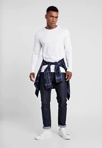 Witte lange mouwen shirt, donkere spijkerbroek, marineblauw geruit shirt om de taille geknoopt, en witte sneakers. Casual outfit met contrasterende kleuren.