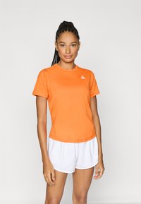 Πορτοκαλί αθλητική μπλούζα με στρογγυλή λαιμόκοψη και κοντά μανίκια, κατασκευασμένη από ελαφρύ υλικό, σε συνδυασμό με λευκό αθλητικό σορτς.