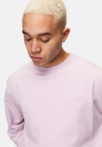 Ljusrosa sweatshirt med en texturerad, broderad logotyp på bröstet. Har rund hals och ribbade ärmslut. Mjuk tygkvalitet, avslappnad design.