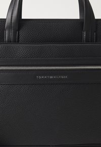 Black leather handbag with a textured finish, featuring dual handles, a zip closure, and a silver logo detailing on the front.