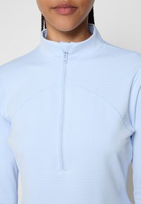 Maglione azzurro chiaro con zip, caratterizzato da un colletto alto, tessuto testurizzato e una vestibilità aderente con cuciture visibili lungo le cuciture.