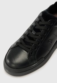Sapatilha de couro preto com acabamento liso, forro de tecido texturizado, seis ilhós pretos e atacadores pretos. Apresenta um logótipo em relevo na lateral.
