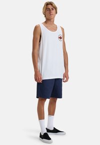 White cotton tank top with red and brown mountain logo, paired with navy shorts. Model wears white socks and black slip-on shoes.