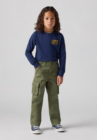 Camicia a maniche lunghe blu navy con logo giallo; pantaloni cargo verde oliva con tasche laterali; sneakers blu con accenti bianchi; design dritto.