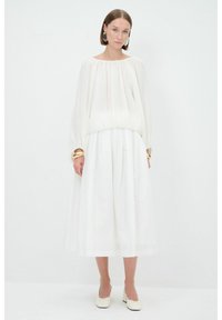 Weiße, locker sitzende Bluse mit puffärmel, gerafftem Detail in der Taille, kombiniert mit einem voluminösen, bodenlangen weißen Rock und minimalistischen Schuhen.