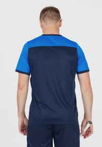 Mies, der in marineblauer und blauer Sportkleidung gekleidet ist und einen Tennis-Schläger mit weißem Griff hält, steht mit dem Rücken zur Kamera vor einem weißen Hintergrund.