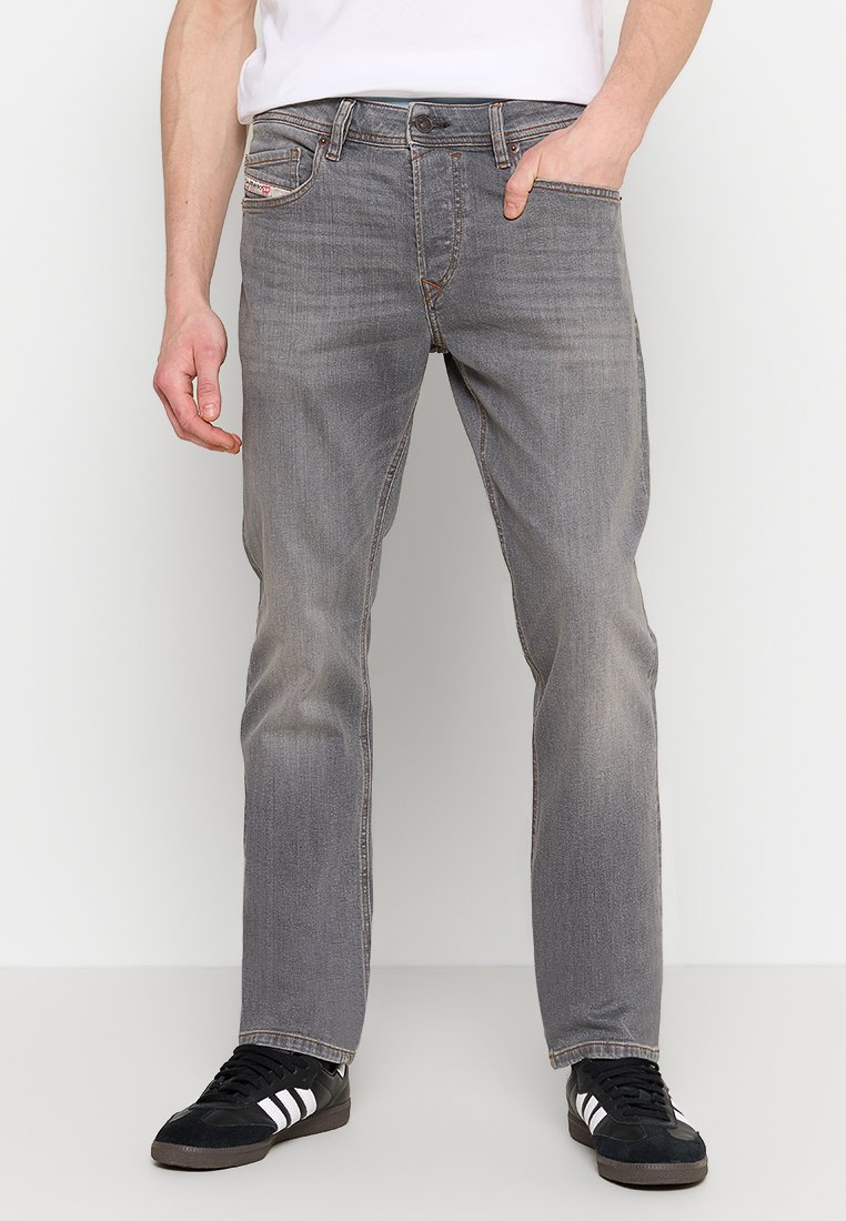 Man in vervaagde grijze jeans en zwarte sneakers met witte strepen, staand met een hand in de zak tegen een witte achtergrond.