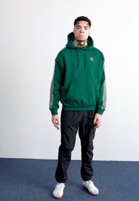 Sweatshirt verde com três riscas brancas nas mangas, combinado com calças pretas e ténis brancos, de pé contra um fundo liso.