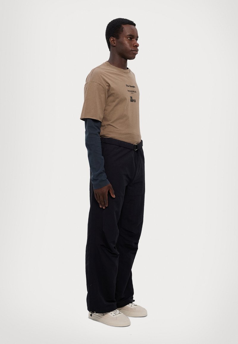 T-shirt di cotone marrone sovrapposta a una maglietta a maniche lunghe blu navy, abbinata a pantaloni neri e sneaker beige, in piedi contro uno sfondo chiaro.