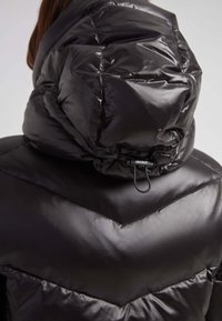 Czarna puchowa kurtka o błyszczące, pikowanej powierzchni. Posiada wysoki kołnierz, kaptur ze sznurkiem oraz wzór w zygzaki na korpusie.