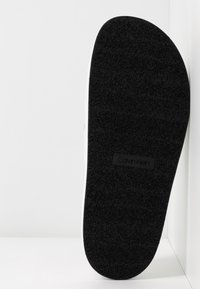 Semelle de sneaker en caoutchouc noir avec une finition texturée. Présente un bord blanc et une étiquette noire avec "Calvin Klein" en relief.