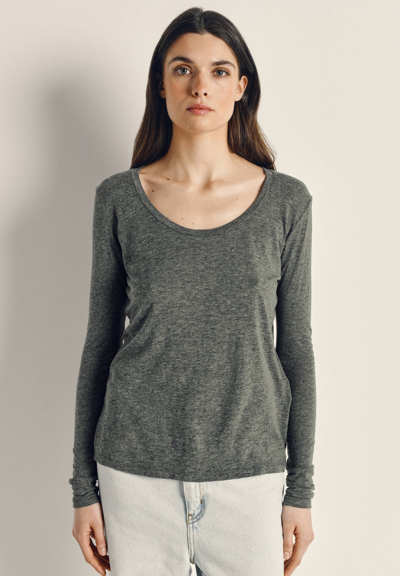 Kvinna med långt brunt hår som bär en grå långärmad topp med rund halsringning och ljusa byxor, stående mot en enkel ljus bakgrund.