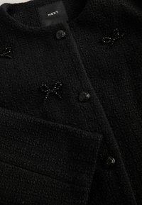 Black textured coat featuring a round neck, two front buttons, and decorative bead accents in the shape of bows. Brand label visible inside.