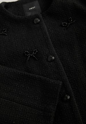 Black textured coat featuring a round neck, two front buttons, and decorative bead accents in the shape of bows. Brand label visible inside.