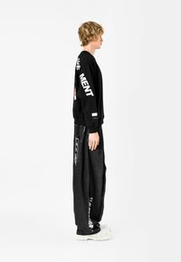 Sudadera negra con texto blanco en las mangas, combinada con pantalones sueltos de color gris oscuro que tienen gráficos laterales. El modelo lleva zapatillas de plataforma blancas.