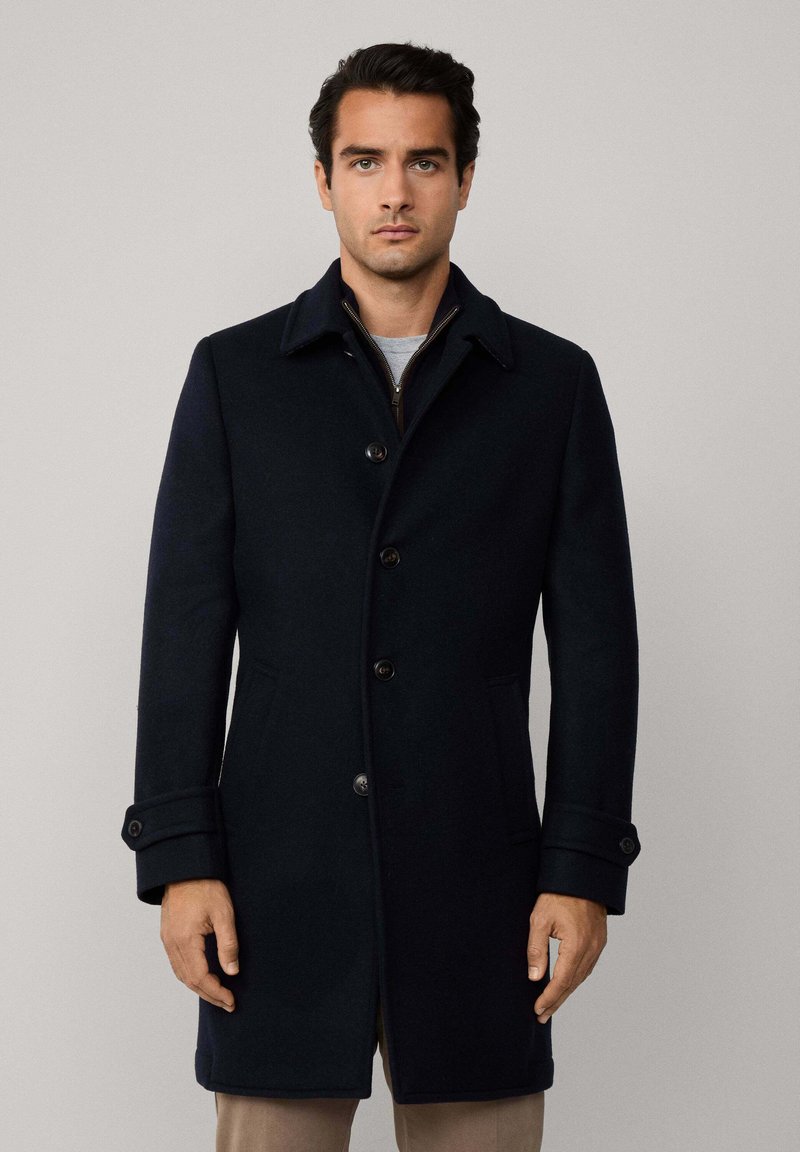 Uomo che indossa un soprabito nero con bottoni e polsini abbottonati, in piedi contro uno sfondo grigio chiaro, rivolto verso l'avanti.