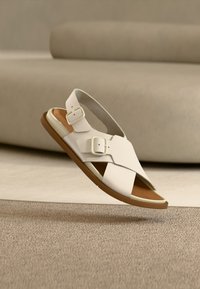 Άσπρο σανδάλι κατασκευασμένο από λείο υλικό, με δύο ρυθμιζόμενα λουριά και ανοιχτό καφέ τακούνι. Ανοιχτός σχεδιασμός με σταυρωτά λουριά.