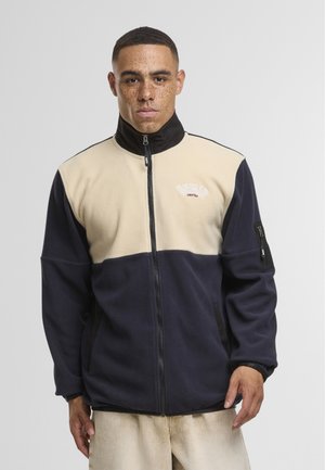 Young man with short hair and freckles wearing a beige and navy zip-up fleece jacket with "UNFAIR" text on the chest.