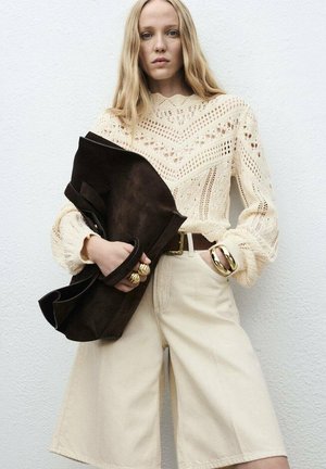 Pull en tricot crème avec un motif ajouré, shorts beige clair, et un sac en daim brun foncé. Des bracelets en or accentuent le look.