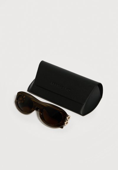 Γυαλιά ηλίου με σχέδιο χελώνας και σκουρόχρωμους φακούς δίπλα σε μαύρη θήκη με υφή σε λευκό φόντο.