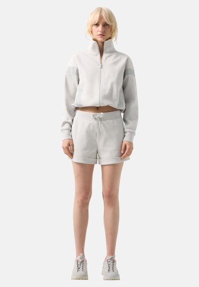 Jeune femme aux cheveux blonds portant une veste zippée gris clair, un short à cordon assorti et des baskets blanches, debout, sur un fond blanc.