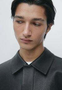 Sort læderjakke med krave, som har en glat tekstur og et struktureret design. Diskrete detaljer omkring knapområdet.