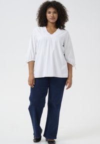 Weißes Blusenshirt mit vertikalen Streifen, V-Ausschnitt, Rüschensaum und Dreiviertelärmeln. Kombiniert mit dunkelblauen weiten Jeans und schwarzen Schuhen.