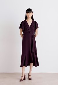 Rochie draped de culoare mov închis, cu mâneci scurte cu volane, talie legată și tiv evazat. Purtați cu tocuri cărămizii, pe un fundal deschis.