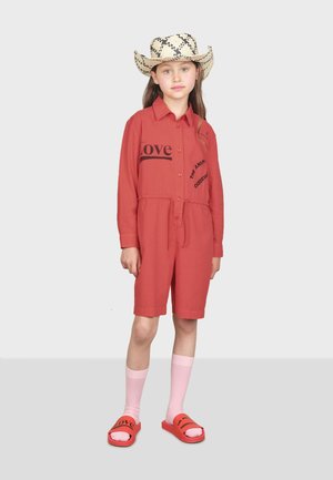 Rompers en coton rouge avec des manches courtes et une taille à cordon, portant un texte noir. Assortis avec des chaussettes rose jusqu'aux genoux et des sandales rouges à enfiler.