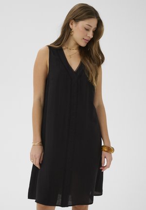 Femme aux longs cheveux bruns regardant vers le bas, portant une robe noire sans manches à col en V et des bijoux dorés, debout devant un fond clair uni.