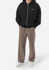 Svart zip-up hoodie med framfickor och logotyp, parat med ljusbruna veckade byxor och vita sneakers. Enkel, avslappnad design.