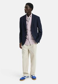Blazer blu navy sopra una camicia a righe rosa e bianche, pantaloni crema e sneakers blu con accenti bianchi. Focus su vestibilità sartoriale e colori a contrasto.