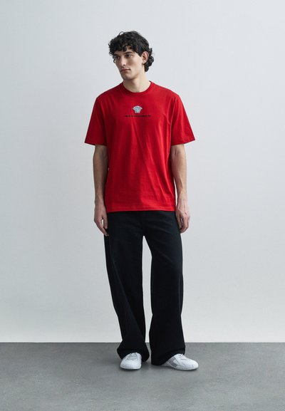 Κόκκινο βαμβακερό t-shirt με κοντά μανίκια και στρογγυλή λαιμόκοψη, με κεντημένο γραφικό σχέδιο στο στήθος. Φορεμένο με μαύρο παντελόνι φαρδύ στις γάμπες και λευκά αθλητικά παπούτσια.