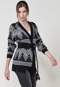 Cardigan a motivo bianco e nero con colletto a scialle, orlo frangiato e cintura annodata. Realizzato in materiale a maglia con maniche lunghe.