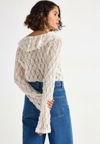 Donna con capelli ricci che indossa un top lungo a maniche trasparenti in pizzo bianco e jeans blu a vita alta, in piedi con le mani dietro la schiena.
