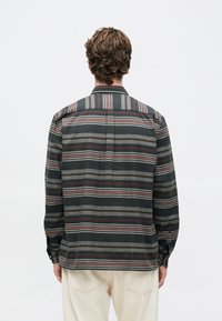 Mężczyzna z kręconymi włosami ubrany w koszulę z długim rękawem w paski w ciemnych i stłumionych odcieniach czerwieni, szarości i beżu, zestawioną z jasnymi spodniami, odwrócony plecami.