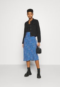 Giubbotto di denim nero, top nero, gonna midi a fantasia blu, stivali militari neri, borsa nera piccola, in piedi contro uno sfondo chiaro.