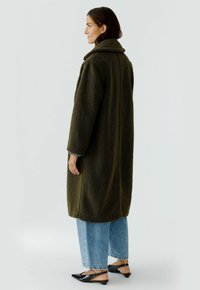 Grün, flauschiger, übergroßer Mantel mit Kragen, kombiniert mit weit geschnittenen hellblauen Jeans und spitzen schwarzen Ballerinas. Weicher Stoff und lange Ärmel.
