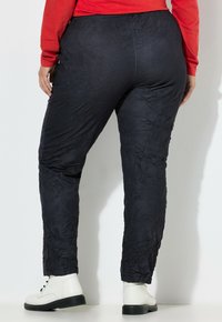 Person, die zerknitterte schwarze Hose, weiße knöchelhohe Stiefel und ein rotes langärmliges Oberteil trägt, steht vor einem schlichten hellen Hintergrund.