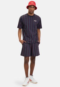 Camicia a maniche corte a righe navy e pantaloni corti coordinati con dettagli rossi, abbinati a un cappello a secchiello rosso e scarpe da ginnastica bianche.