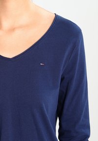 Laivastonsininen pitkähihainen paita, jossa on V-kaula-aukko, valmistettu pehmeästä kankaasta. Rintamuksessa pieni brodeerattu logo punaisena, valkoisena ja sinisenä.