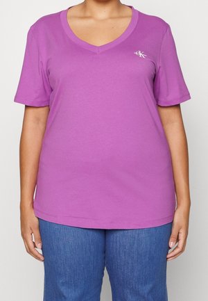 Lila T-Shirt mit kurzem Ärmel und V-Ausschnitt aus weicher Baumwolle. Verfügt über ein dezentes Logo auf der Brust und einen legeren Schnitt, kombiniert mit blauem Denim.