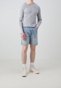 Persona de pie con una camiseta de manga larga de canalé gris claro, pantalones cortos de mezclilla desgastados, calcetines blancos y zapatillas altas beige.