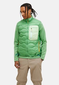 Grüne, isolierte Weste mit einem gesteppten Muster, Reißverschluss-Brusttasche und kontrastierenden orangenen Beschlägen. Leichtgewichtig, mit verstellbarem Saum.