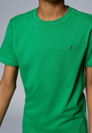 Person iført en ensfarvet grøn T-shirt med korte ærmer og rund halsudskæring med et lille rektangulært logo på venstre bryst.