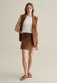 Braunes maßgeschneidertes Weste über einem weißen gehäkelten Oberteil, kombiniert mit einem passenden braunen Rock und geschnallten braunen Sandalen. Glattes Material, minimalistisches Design.