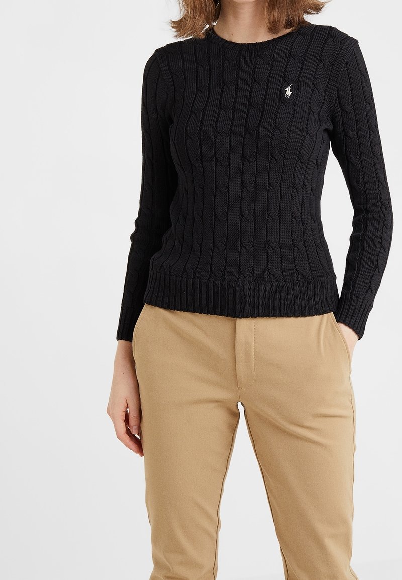 Femme portant un pull en maille torsadée noir à manches longues avec un logo blanc et un pantalon beige, debout avec une main dans la poche.