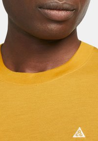 Cotton t-shirt in mustard yellow with a crew neckline. Features a small white logo in the shape of a triangle near the neckline.